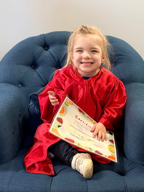 Violette wears a red graduation robe and beams, holding a certificate of graduation from feeding therapy.