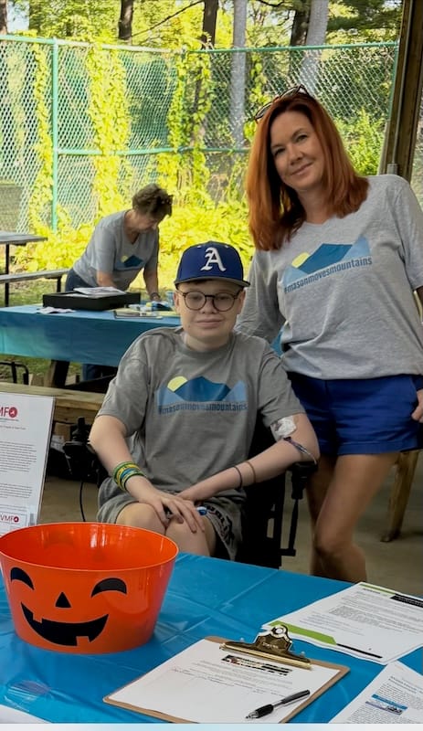 Riley, a white boy with glasses, sits in his wheelchair with his mom Shannon, a white woman with mid-length brown hair.