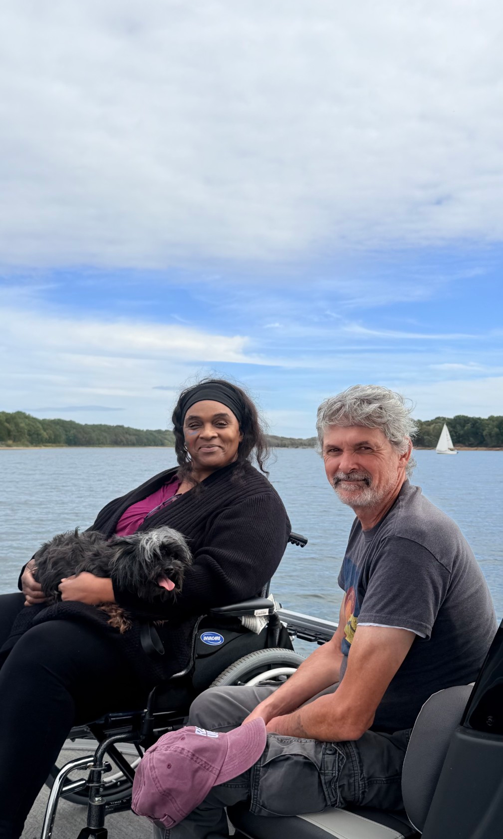 Cheri, a Black woman with long hair, sitting in her wheelchair with a dog in her lap. Her husband, a white man with greying hair, smiles next to her.