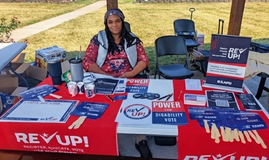 Cheri, a Black woman with long hair, sits at a table of educational materials for disabled voters.