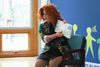 A drag queen seated in a chair in front of a Little Lobbyists background, holding an open book.