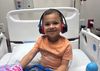 JW, a biracial child with a huge smile, sits in a hospital bed wearing headphones and using a tablet.