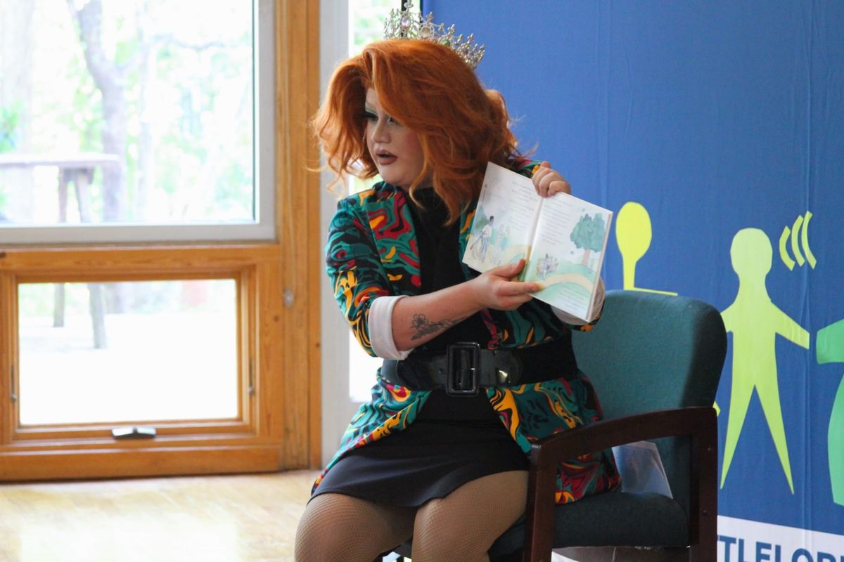 A drag queen seated in a chair in front of a Little Lobbyists background, holding an open book.