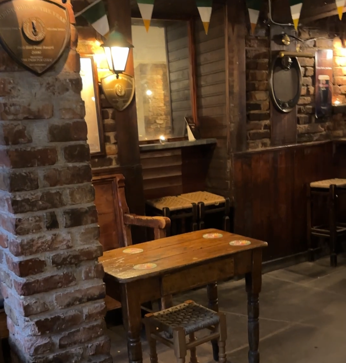 The main barroom at An Spailpín Fánach with wooden tables and stools with woven seats next to exposed brick walls.  A street lamp illuminates the room.  