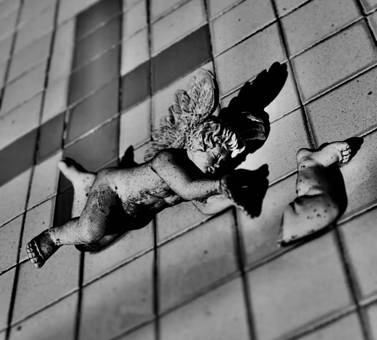 Black and white photo of a tiny cemetery angel, which has fallen on its side. There is third detached angel leg lying next to it.