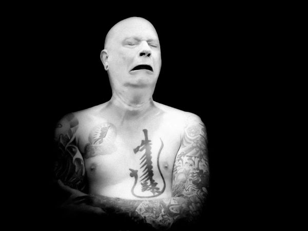 Black and white photo of a bald man, with very white skin, tattooed arms folded across his check. Hie eyes are closed, his mouth downturned, in an exaggerated expression of grief or sorrow. 