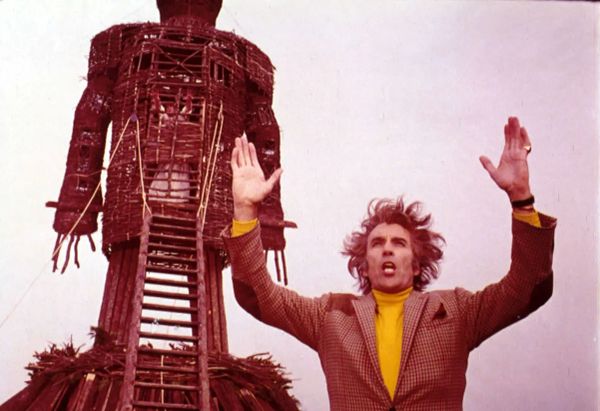 A guy with his arms held open above his head, with flowing hair, and a wild look in his face. Behind him stands a large wicker figure.