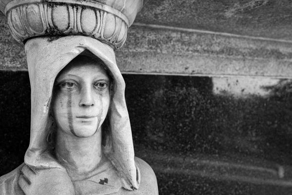 Monochrome, high contrast, close up photo of the face of a cemetery statue of a hooded androgynous figure. Rain stains appear as tear streaks under the eyes.