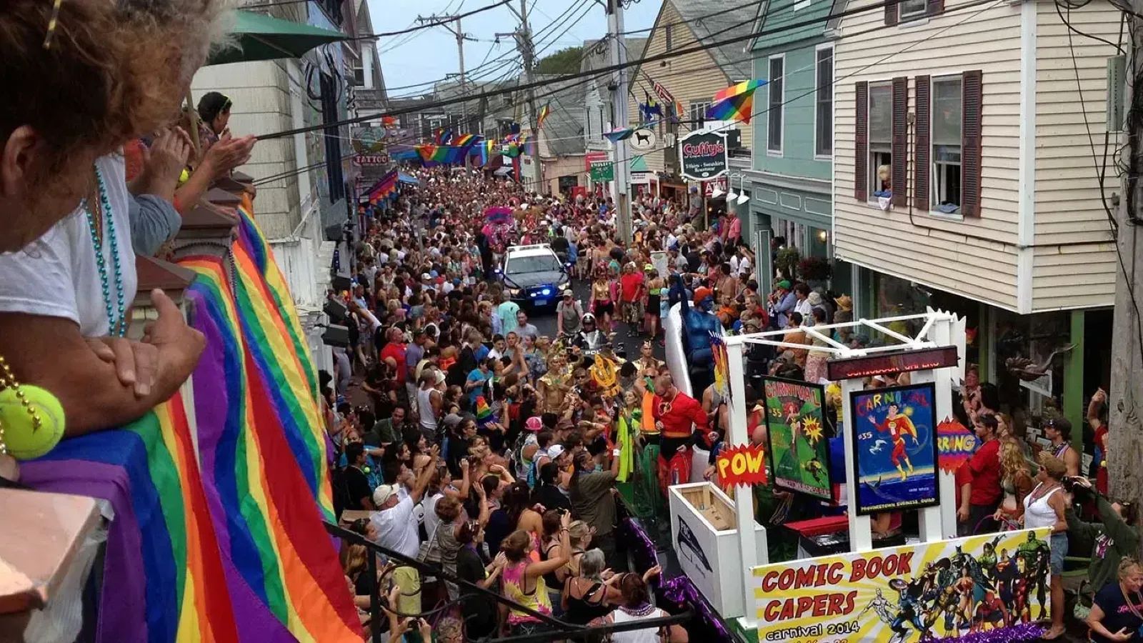 A crowd of people at Provincetown Summer Camp Carnival with a Pride flag hanging from a window