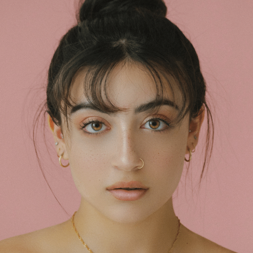 image of a womans face with dark hair, ear rings, nose ring with pink background