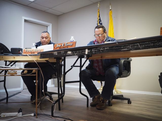 Mayor Peter Nieto and Town Council Member Jose "Richard" Torres listen as Rebecca Anthony made her case for the issuance of alcohol permits for four art openings at La Galería at the Shaffer Hotel. Mayor Nieto is reviewing Anthony's application. Council Member Torres is positioned in front of a US flag and a New Mexico flag.
