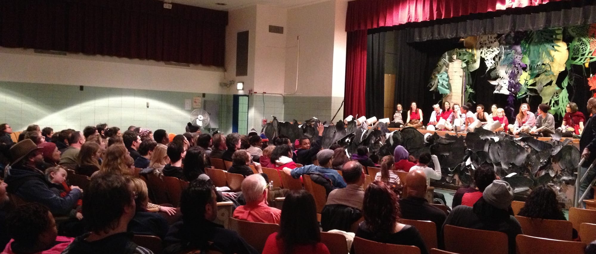 An audience in an elementary school auditorium looking at a stage where actors, both children and adults, are sitting in a Q&A session. The artistic director is standing at the far right.