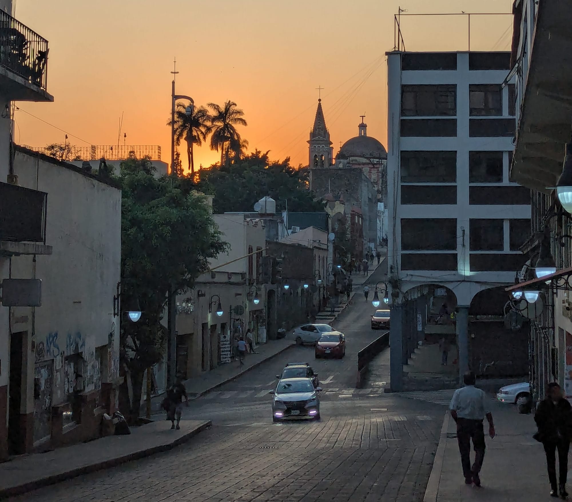 the sun sets behind palm trees next to the dome and spire of the cathedral in Cuernavaca. A street leads up the hill toward the cathedral.