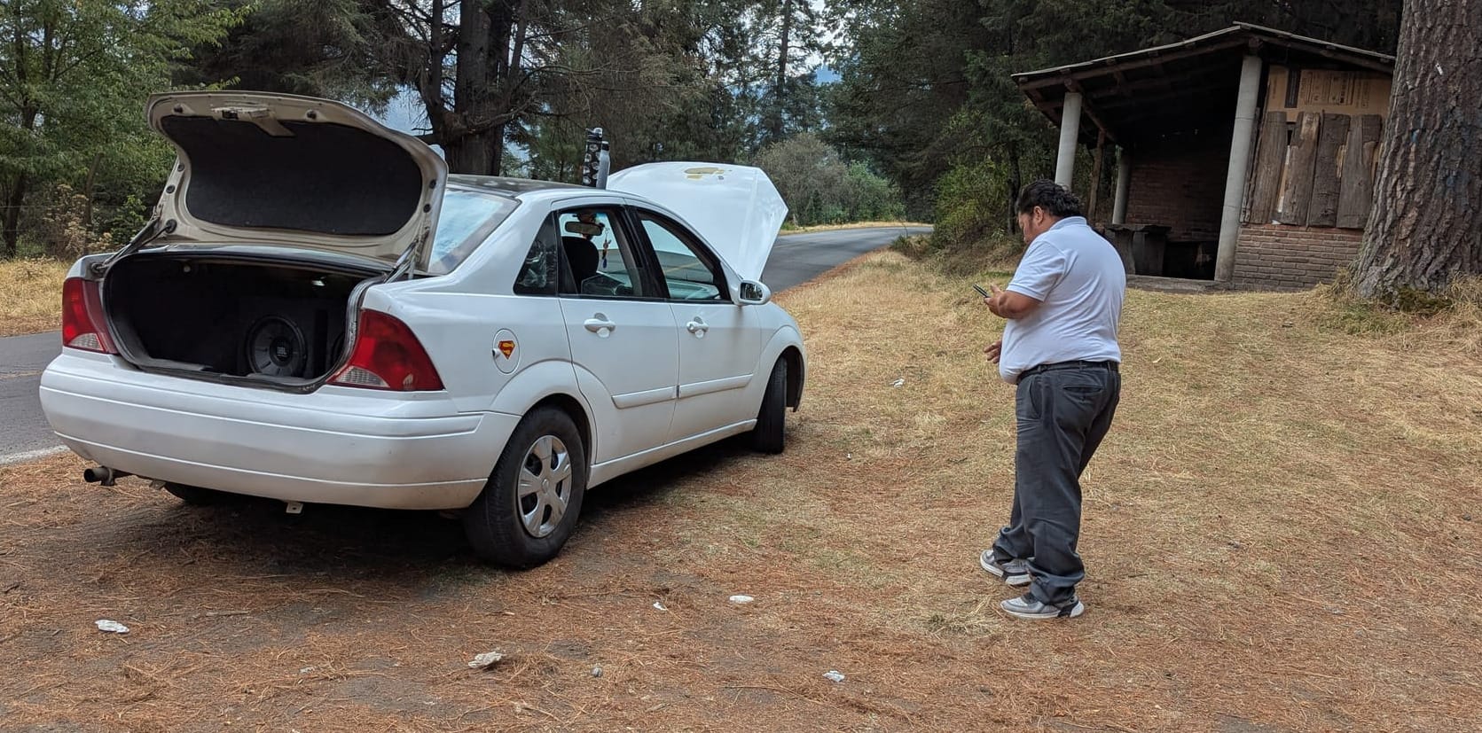 A man looks at his phone at the side of a white car with its hood and trunk open on a mountain road.
