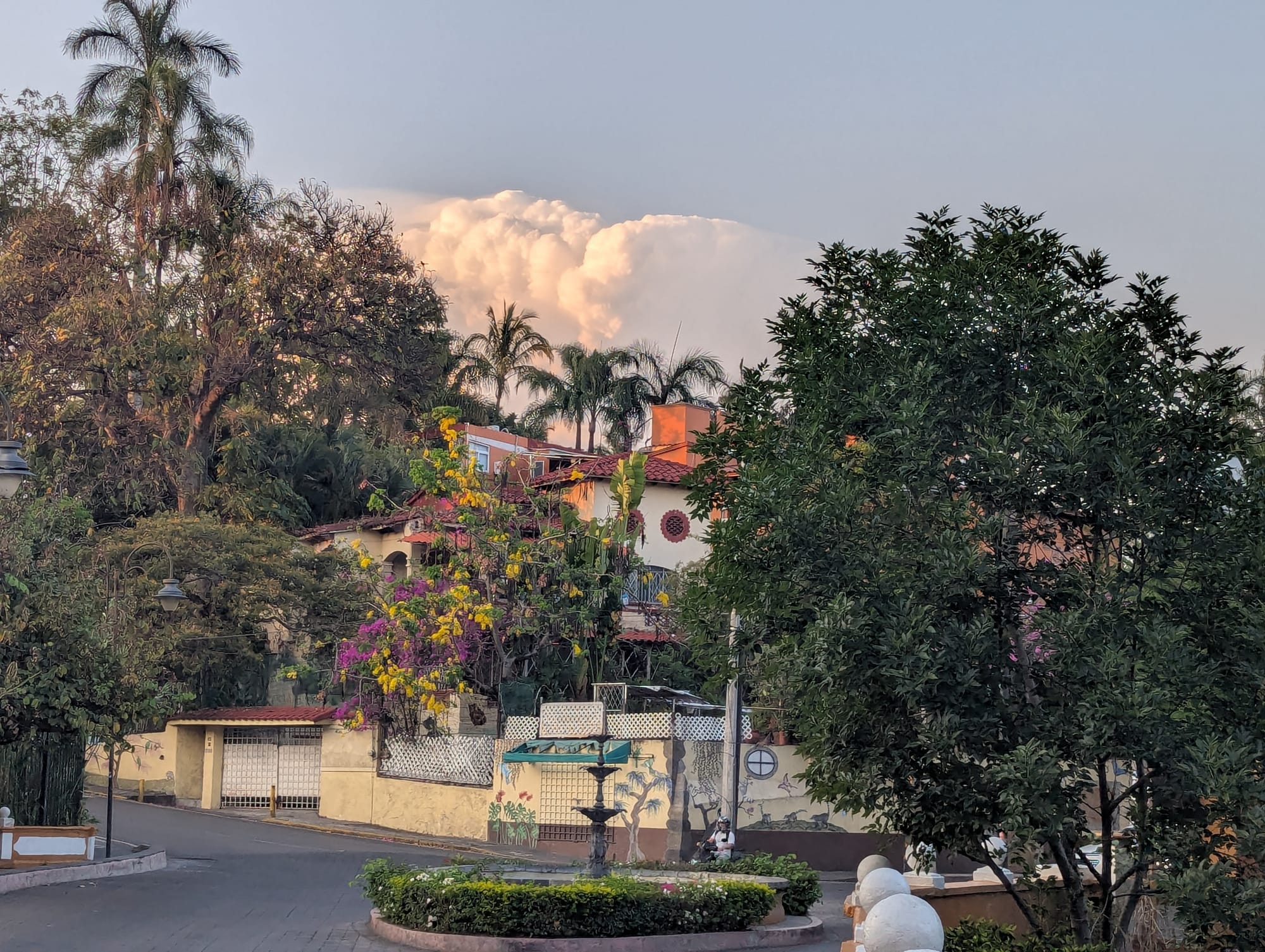 an intersection in Cuernavaca with a fountain and behind it, purple and yellow flowers, a tile-roofed house, and billowing clouds