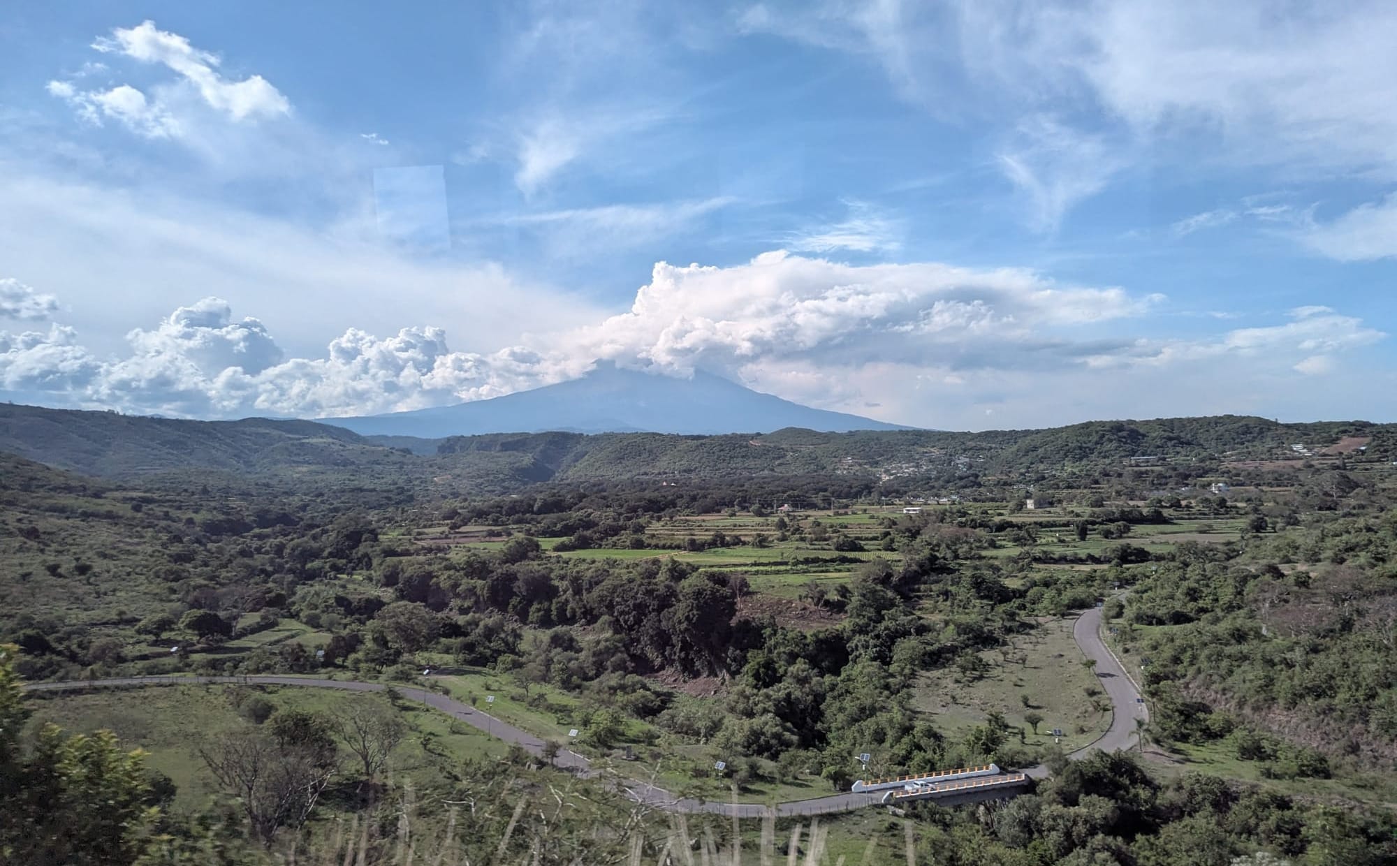 the volcano of Popocatepetl, seen across green fields and woodlands. Smoke is coming out of the volcano's cone and clouds surround it.