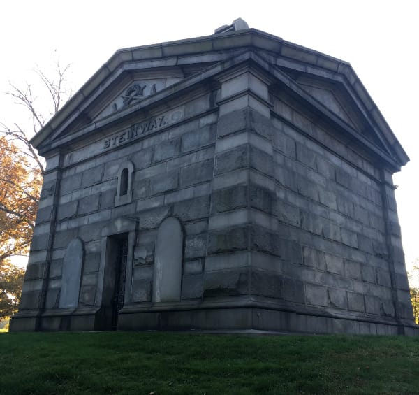 a large gray square mausoleum. the photo is taken looking up from below. The name Steinway appears over the door, below the cornice.
