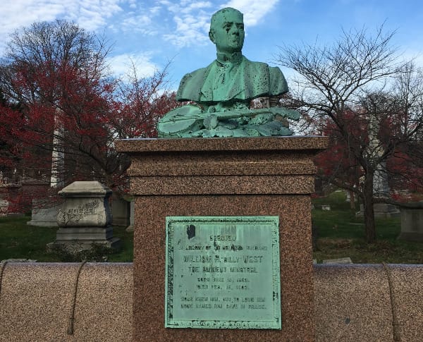 a monument with a green bronze bust of a man, and below it a bronze banjo. A plaque lower on the monument identifies him as William "Billy" West