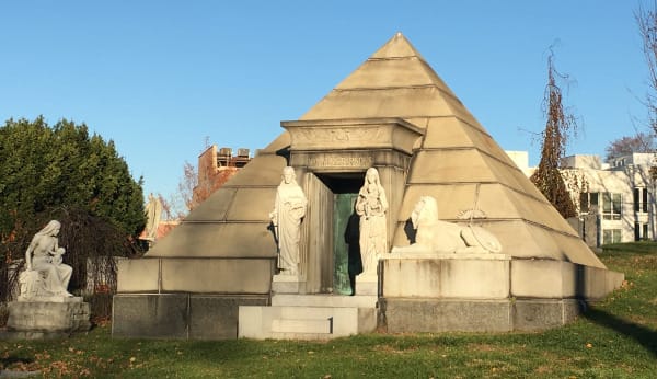 a mausoleum in the shape of a pyramid, with two robed figures on either side of the door and a lion nearby