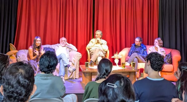 five theater people sitting on a stage in front of a red curtain, while audience members listen