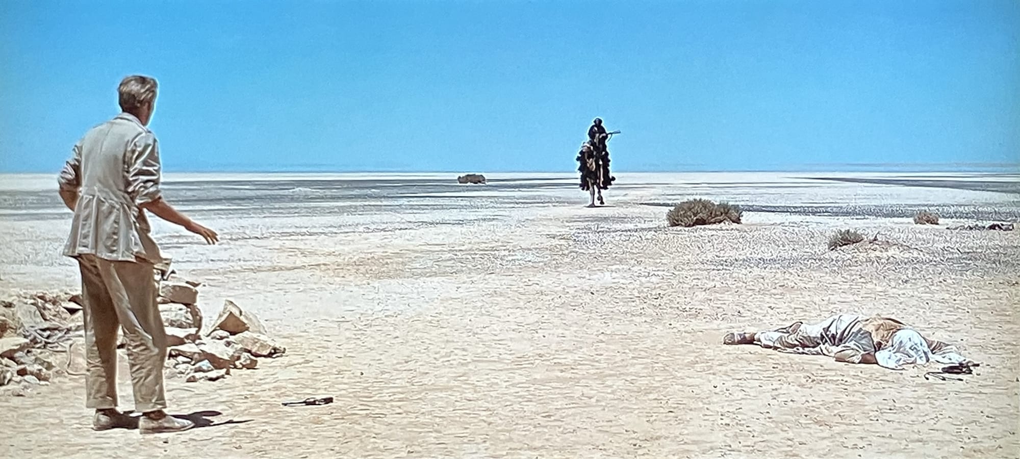 Peter O'Toole, back to the camera, looking at Omar Sharif riding toward him on horseback, with a dead man on the ground