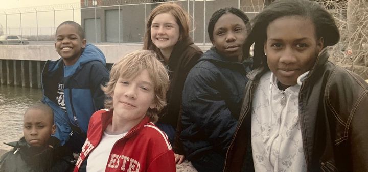 Six young people sitting outside by New York Harbor, looking at the camera for a publicity shot. They are wearing various styles of cold weather coats and clothing.
