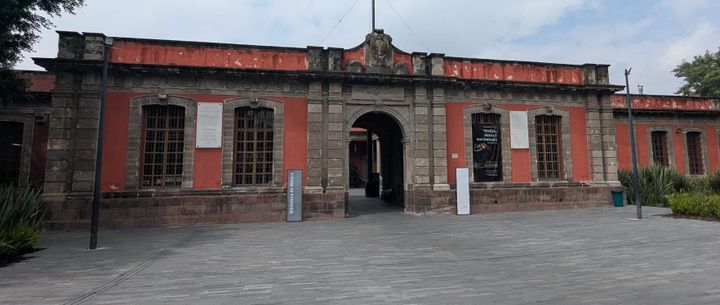 The Library of Mexico, a Spanish colonial building looking like a fortress. It sits back of a wide plaza, with a stone gate flanked by red walls.