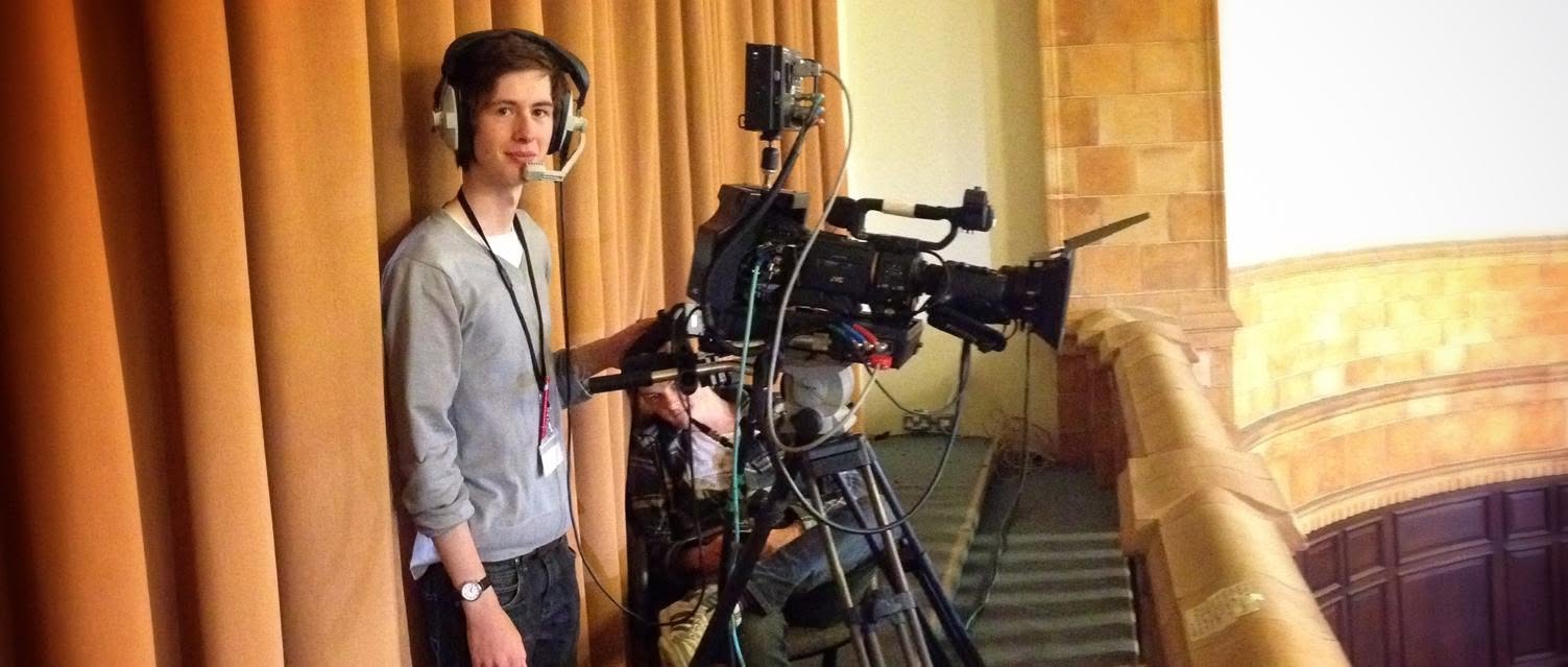Zander Weaver wearing headphones and standing beside a professional film camera in 2012.