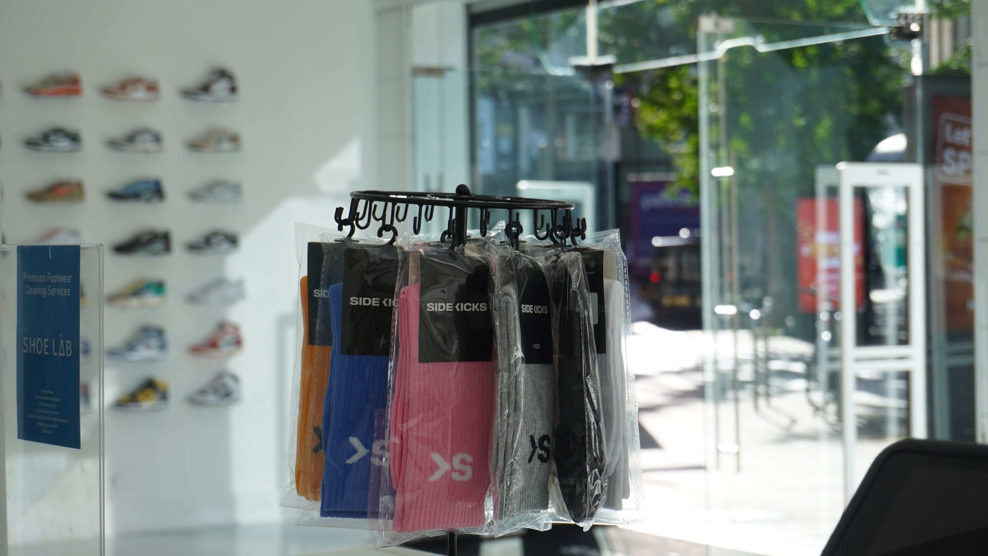 An image of a rack full of colorful crew socks that are branded with the "SK" monogram. The background of the photo is out of focus and shows the storefront. 