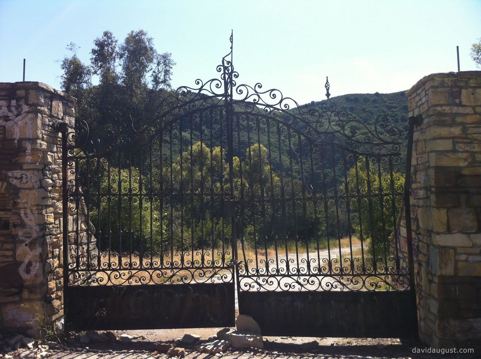 An ornate, misaligned wrought iron gate set between two stone pillars, with a scenic view of rolling hills, a pathway and greenery in the background.