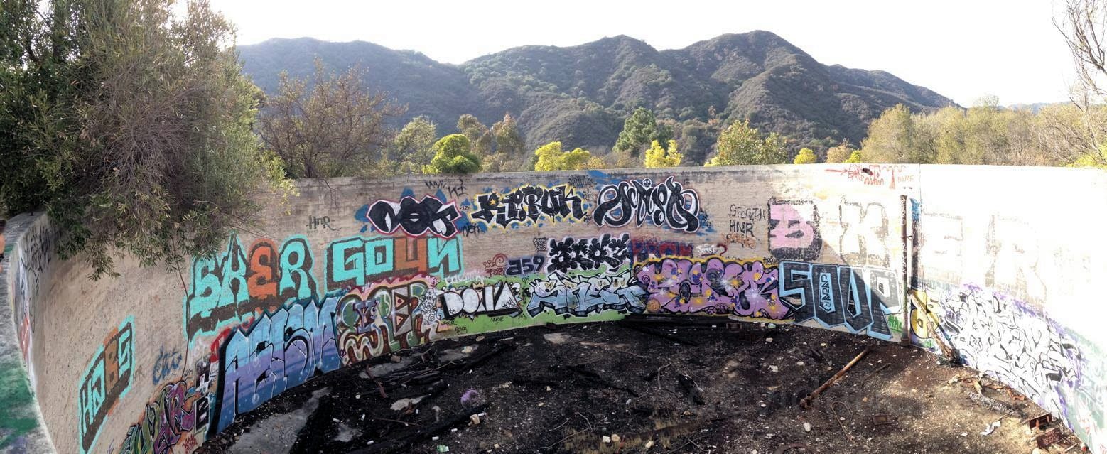An abandoned, graffiti-covered curved concrete structure with a panoramic view of mountains and trees in the background.