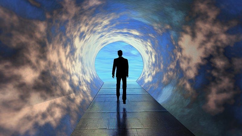 A dark silhouette of a man walks down a cloudy tunnel toward a blue sky.