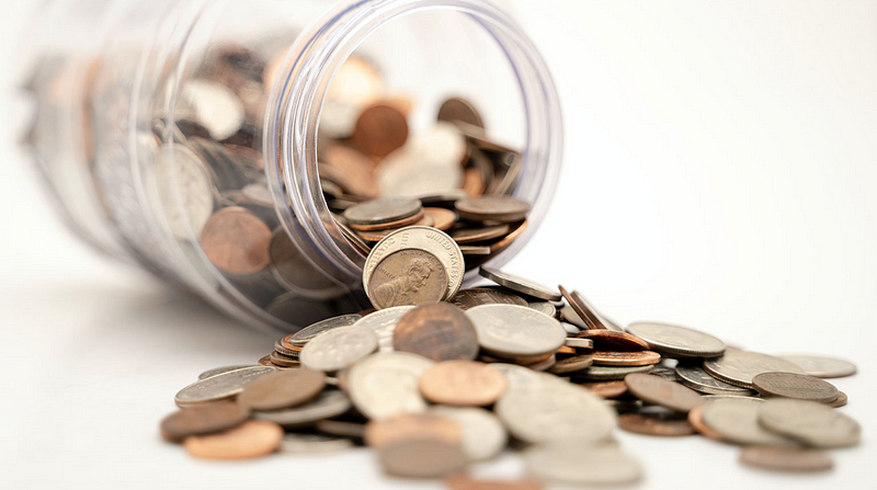 A transparent jar tipped over, with assorted coins spilling out onto a white surface.