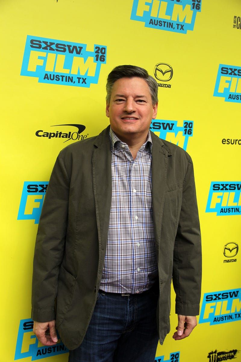 Ted Sarandos standing in front of a primarily yellow step and repeat at SXSW in 2016, he is wearing a brown/greenish jacket,