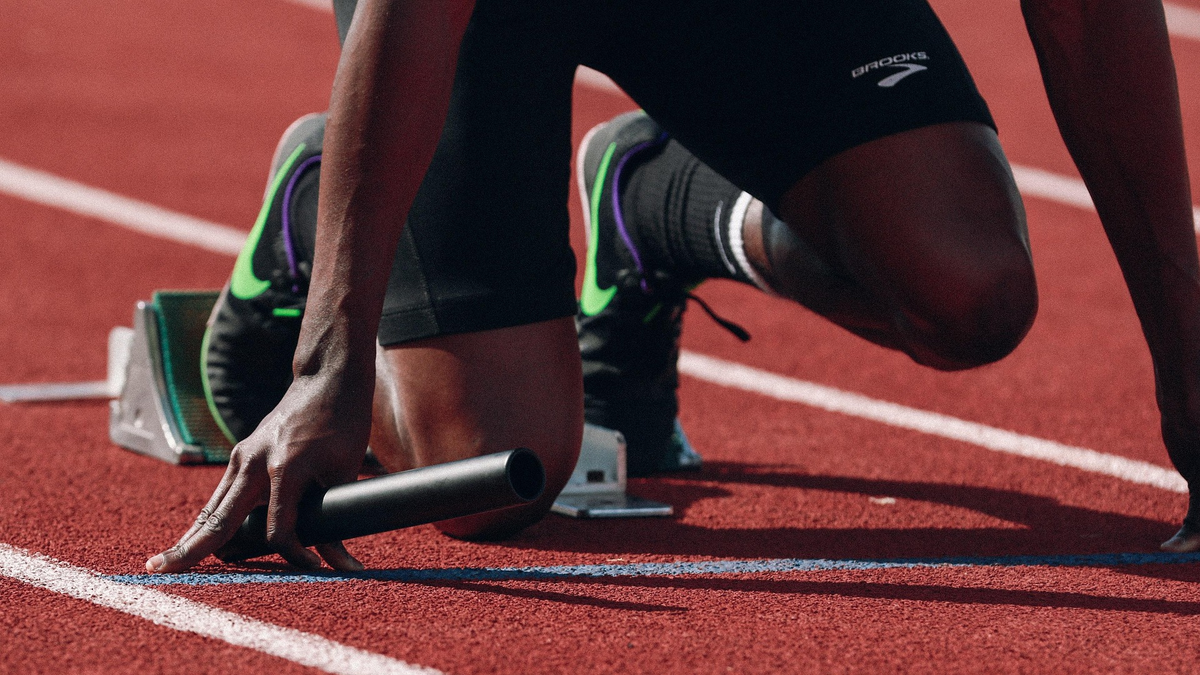 An athlete in a crouched starting position on a red running track, with hands placed on the ground and feet against the starting blocks, ready to sprint.