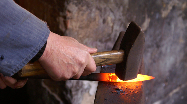 A person's hand holding a hammer striking a hot, glowing piece of metal on an anvil, likely during a blacksmithing process.