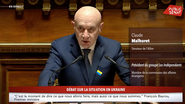 A bald man in a dark blue suit and tie is speaking at a podium in a formal government setting. He has a serious expression an