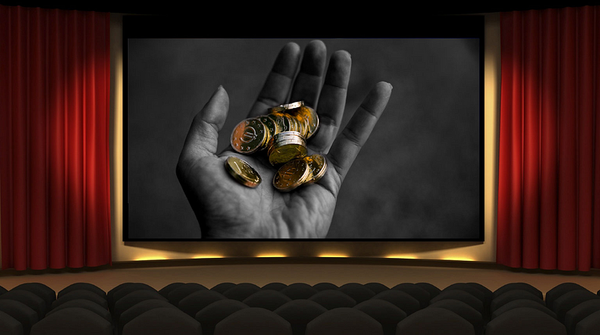 A black-and-white image of a hand holding several coins, displayed on a movie theater screen with red curtains and empty seats in the foreground.
