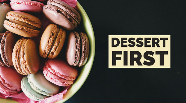 A bowl filled with colorful macarons in various shades of pink, brown, and green, on a dark background. To the right, text overlay reads "DESSERT FIRST."