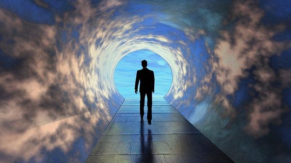 A dark silhouette of a man walks down a cloudy tunnel toward a blue sky.