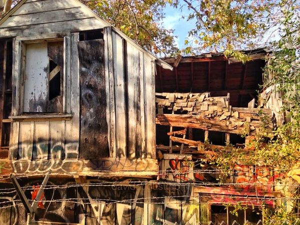 A dilapidated wooden structure with broken walls, windows and roof, surrounded by graffiti and overgrown vegetation, with a barbed wire fence in the foreground.
