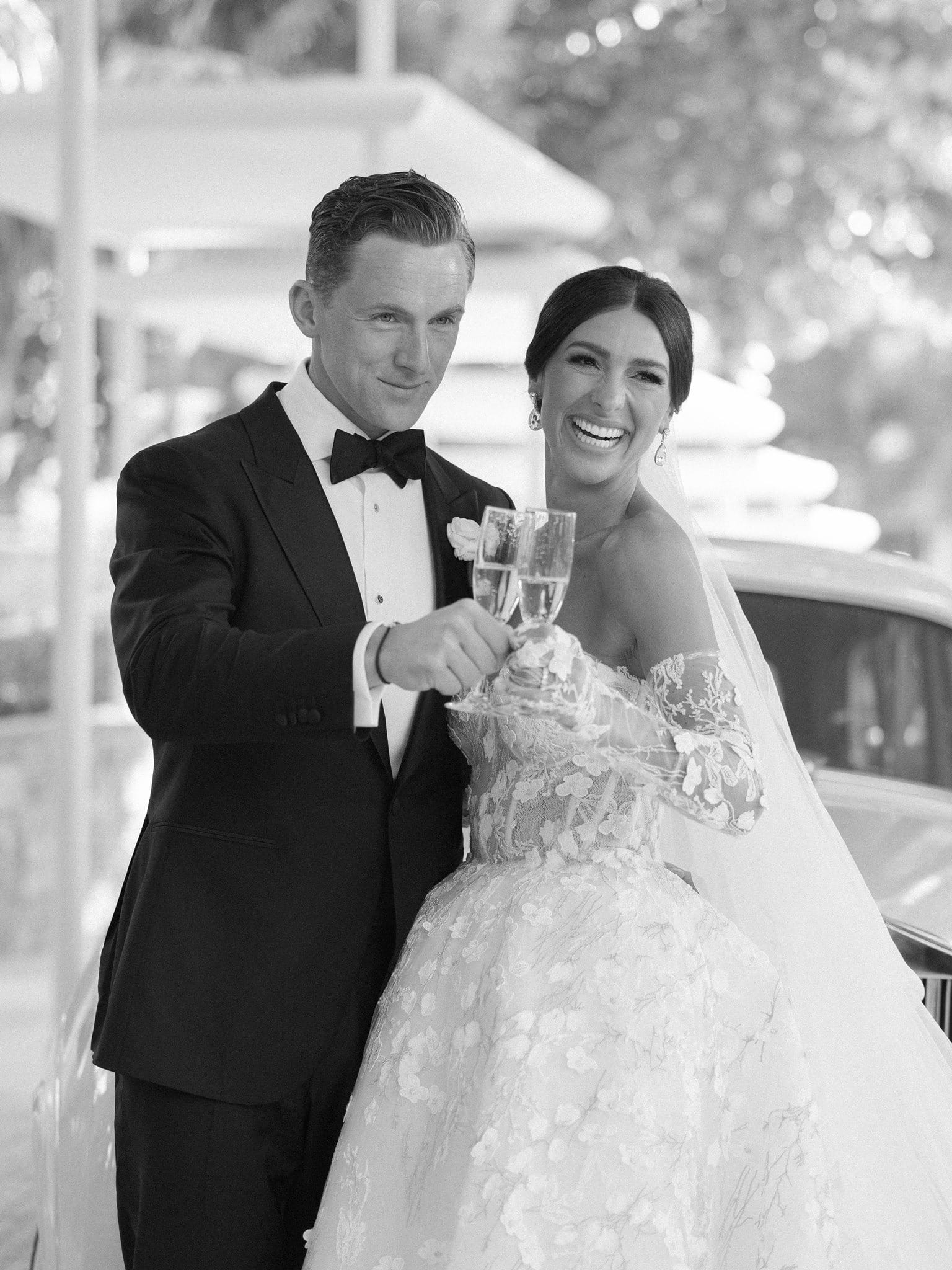 Black and white portrait of a couple toasting with champagne beside a car