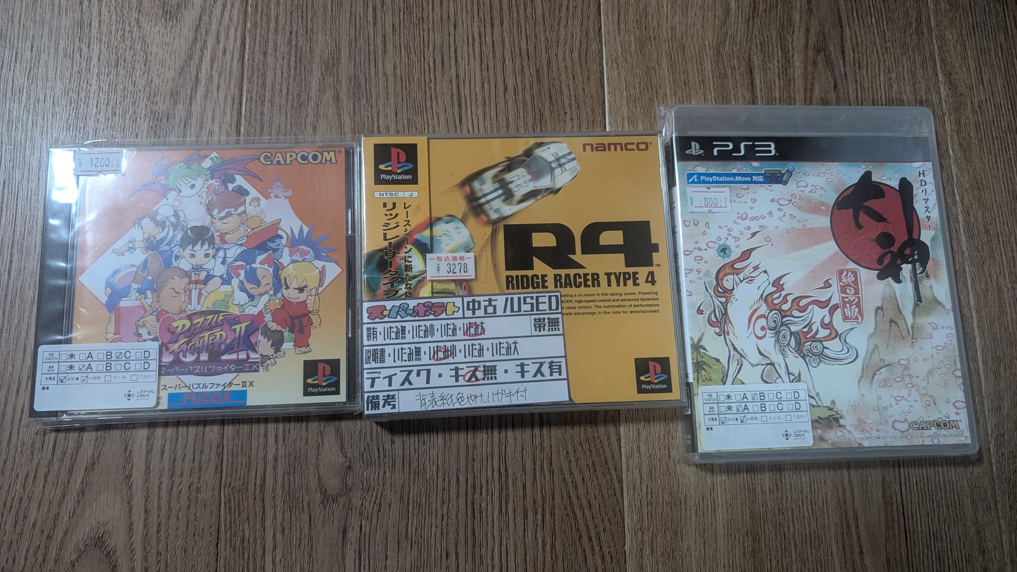 Physical copies of Puzzle Fighter II (PlayStation), R4 Ridge Racer Type 4 (PlayStation), and Okami (PlayStation 3) laying on the ground next to each other.