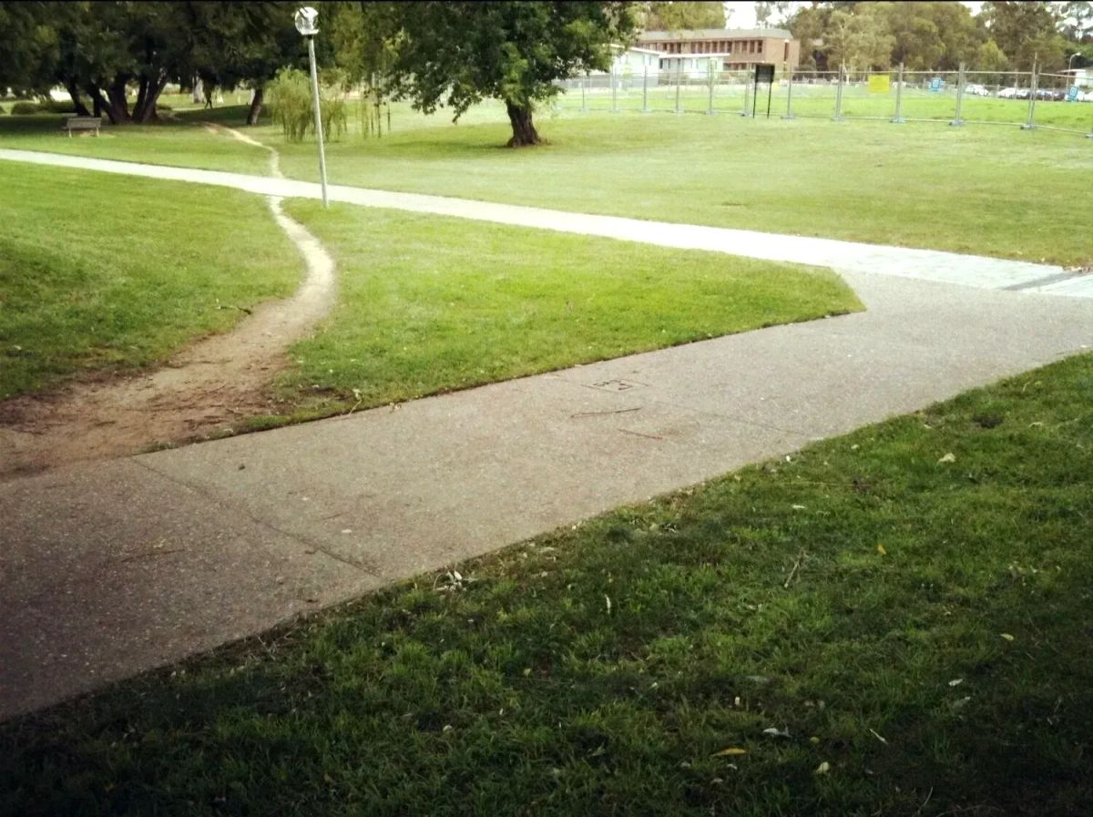 Concrete sidewalk with diagonal dirt path worn into grass, showing user behavior vs. designed infrastructure.