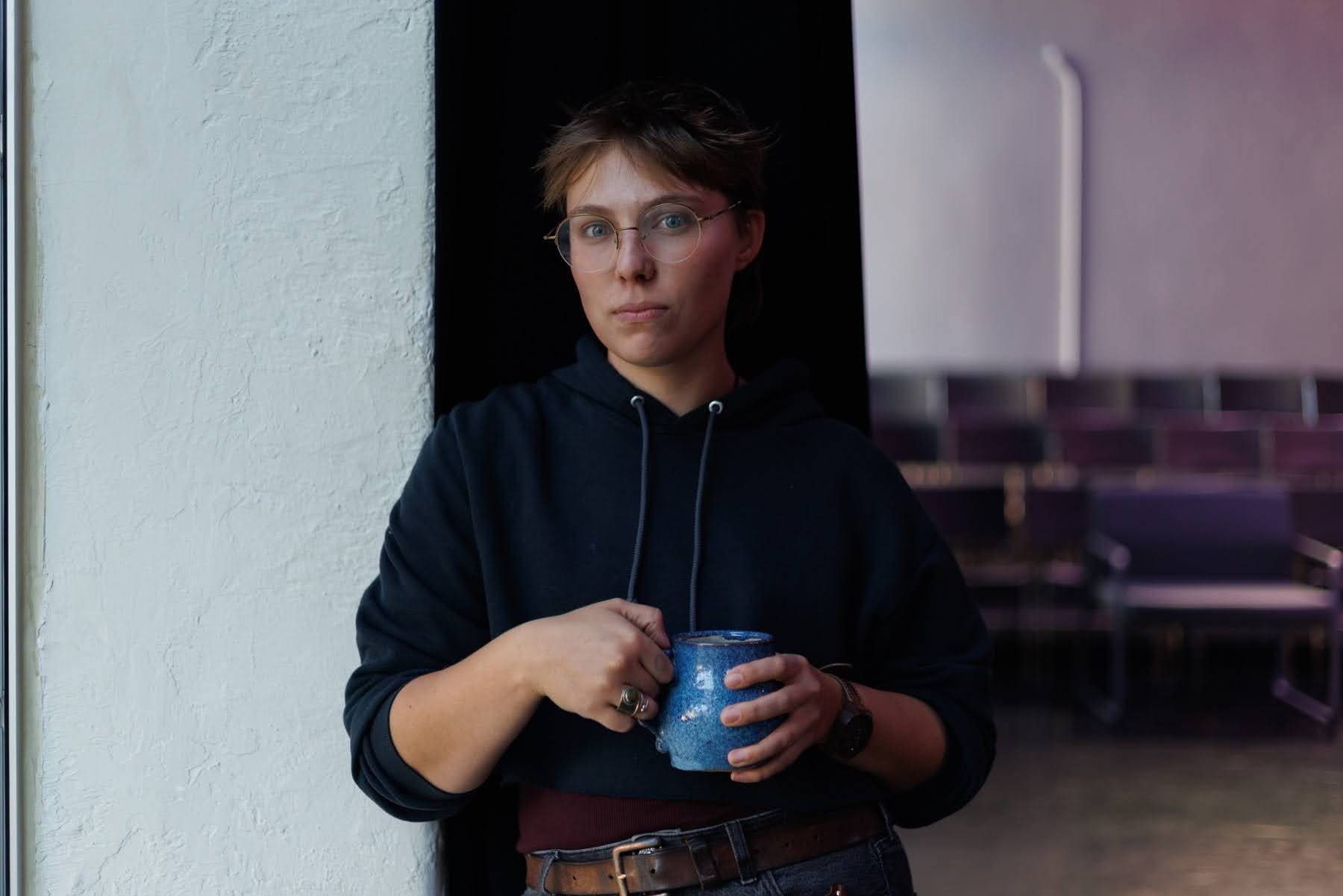 Person wearing glasses and a dark hoodie stands indoors, leaning against a white wall while holding a blue ceramic mug, with empty chairs visible in the background.