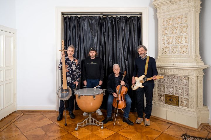 A picture of Arnold Dreyblatt & The Orchestra Of Excited Strings, stood with their instruments against a black curtain.