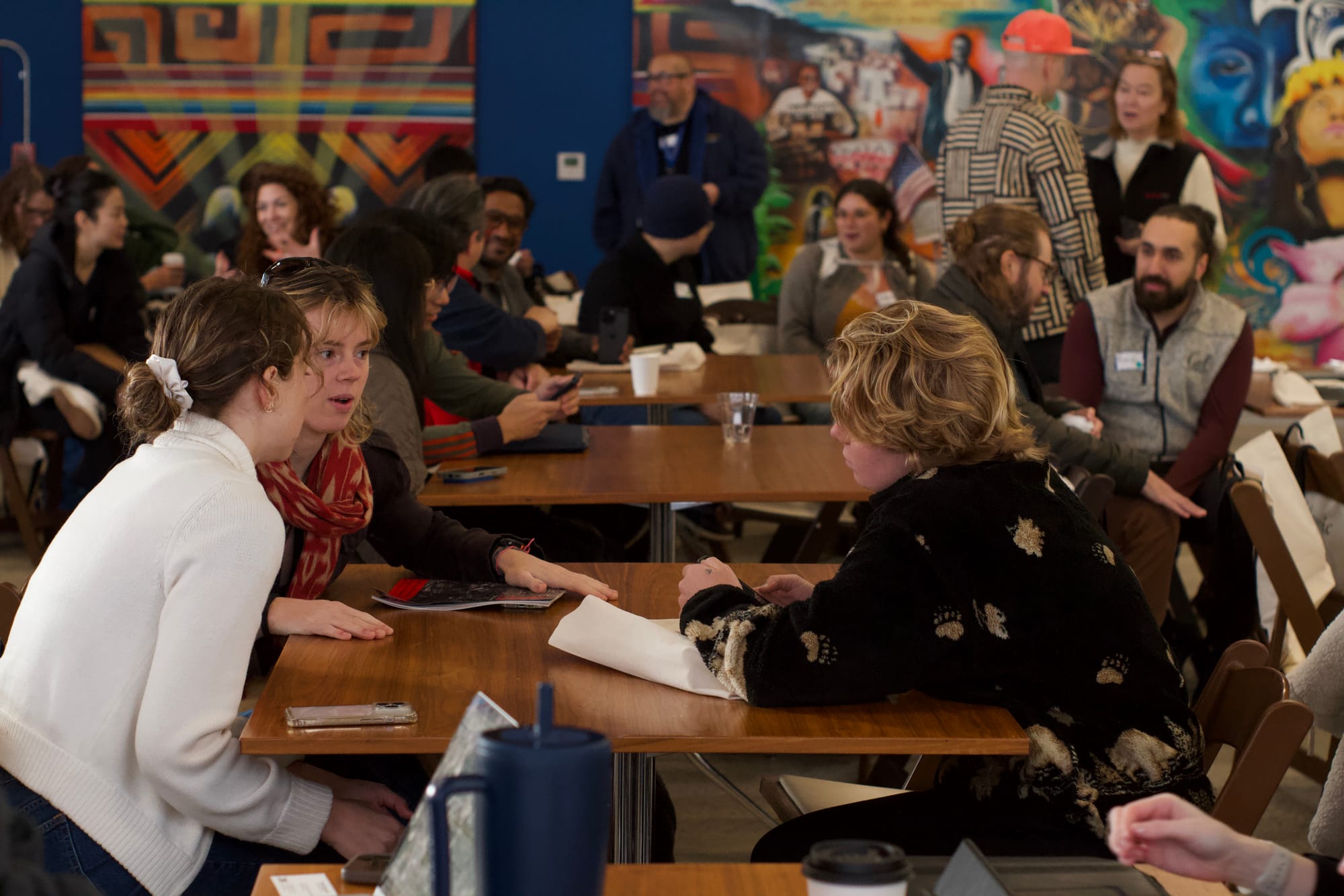 Attendees in small-group conversation at tables during a hackathon, a colorful mural on the wall behind them