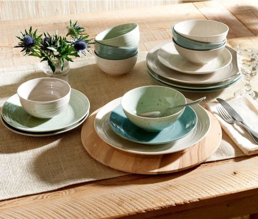 multicolored porcelain dinnerware on the dining table with a burlap table runner; dinner plates, salad plates, dinner bowls, and soup bowls in sage green, blue, gray, and cream