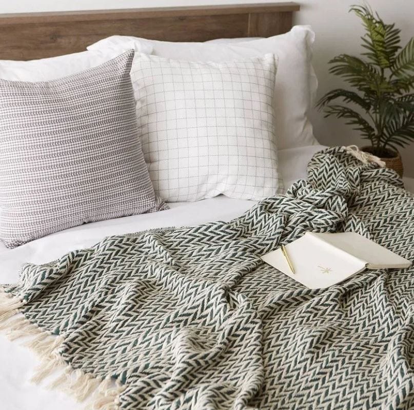 zig zag woven cotton blanket in green and cream on top of a white comforter; two pillows against a wooden headboard and a houseplant in the corner of the room; QVC blankets and throws; Walmart blanket sale