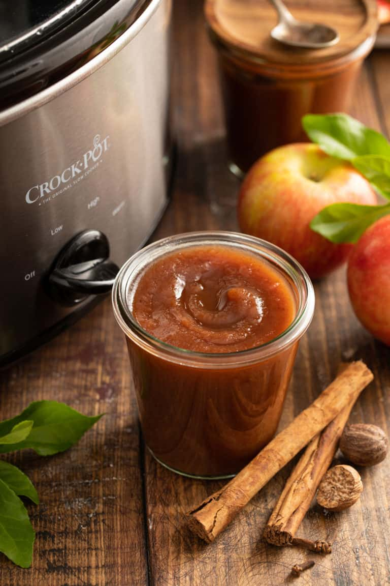 Slow cooker apple butter in a glass jar beside cinnamon sticks, apples, and a crock pot